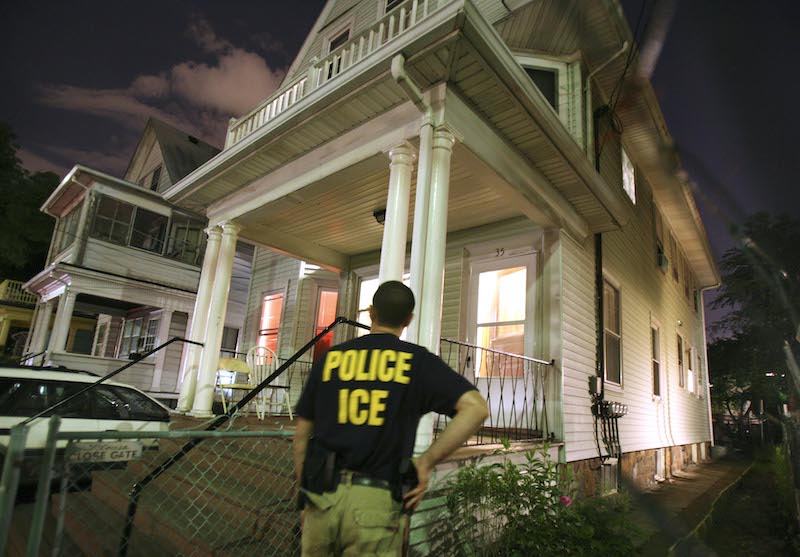 An ICE officer approaches the outside of a house.