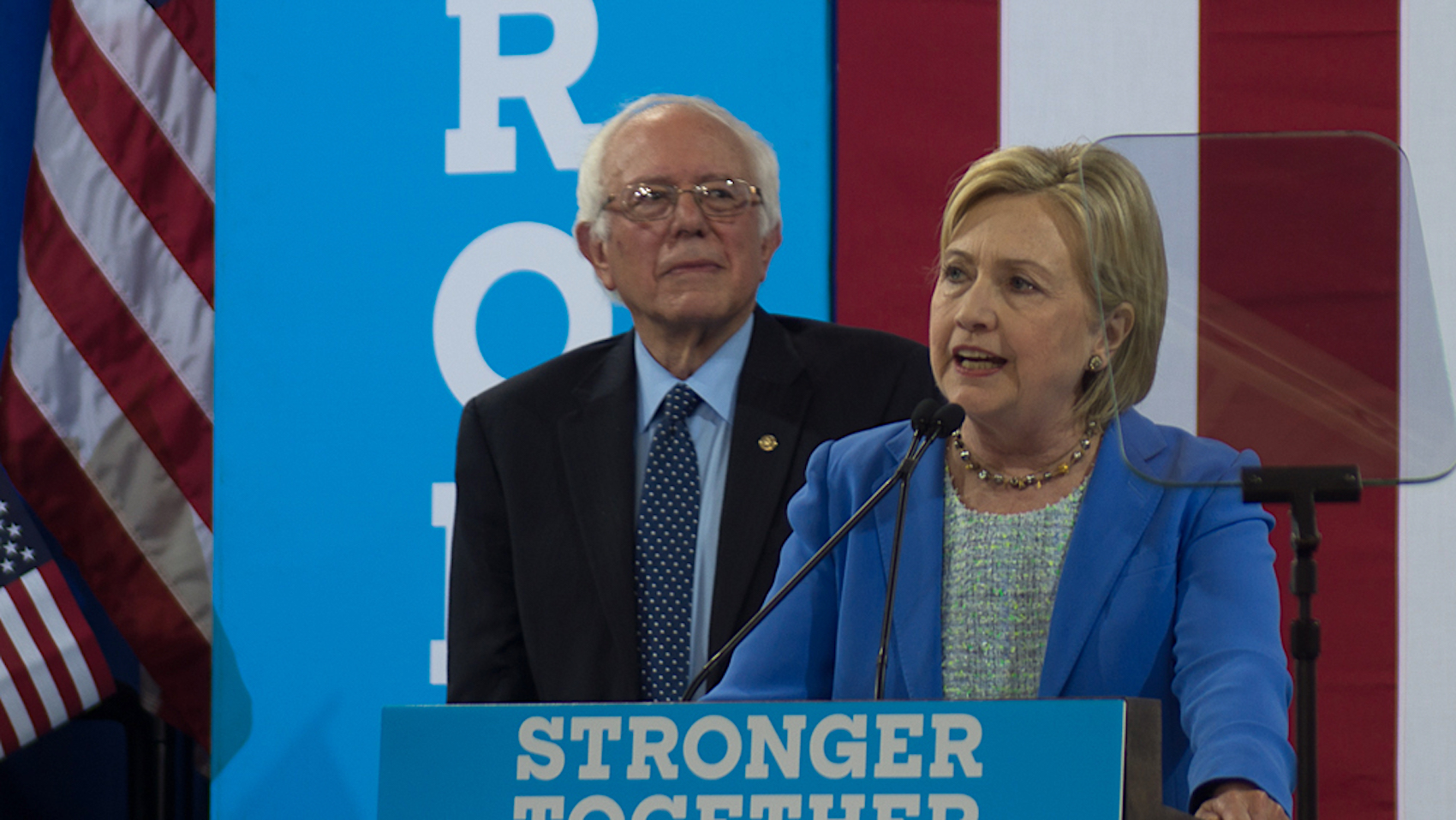 Hillary Clinton speaking at a podium with Bernie Sanders standing behind