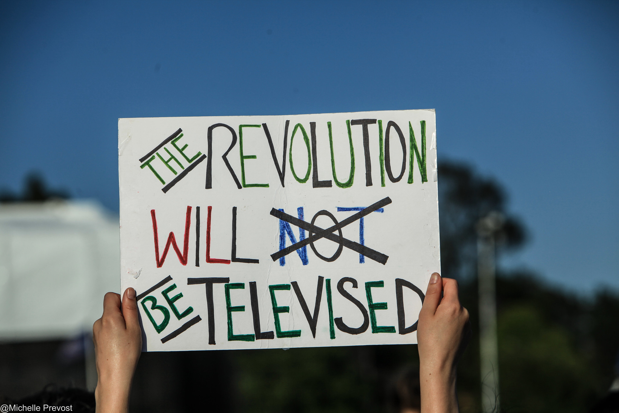 A protest sign reads 'The revolution will not be televised" with the word 'not' crossed out.