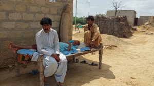 Two men sit on the edges of a cot, one facing the camera. The cot is occupied by a third man stretched out on a blue blanket with his eyes closed and hands folded.