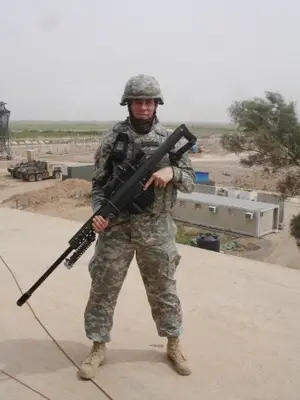 A soldier dressed in fatigues and helmet stands in a desert scene holding a large rifle