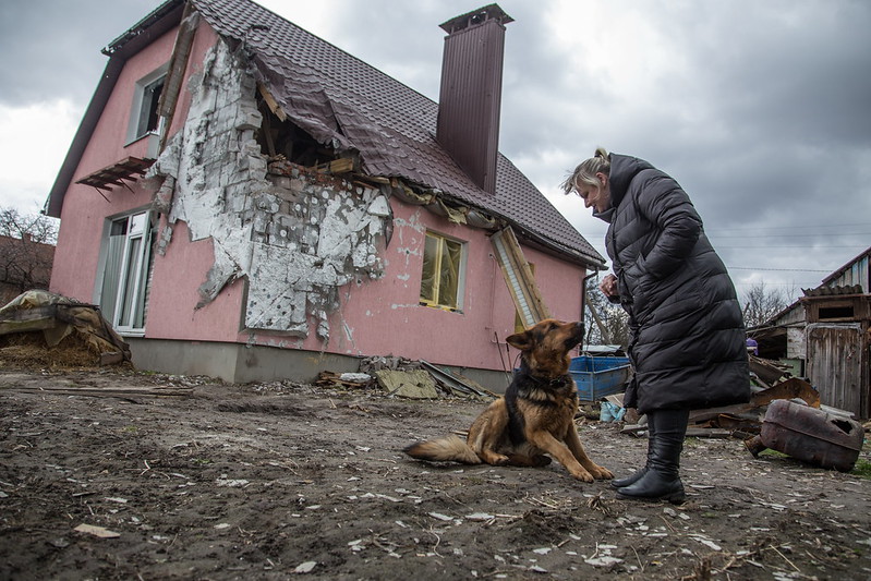 A Ukrainian woman in a winter coat looks down at her large brown and black dog as they stand in front of a small pink house that has been damaged by artillery fire with debris from the destroyed housing surrounding them.