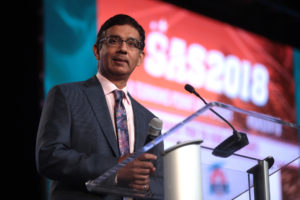 Dinesh D'Souza stands at a podium holding a microphone at a political rally.