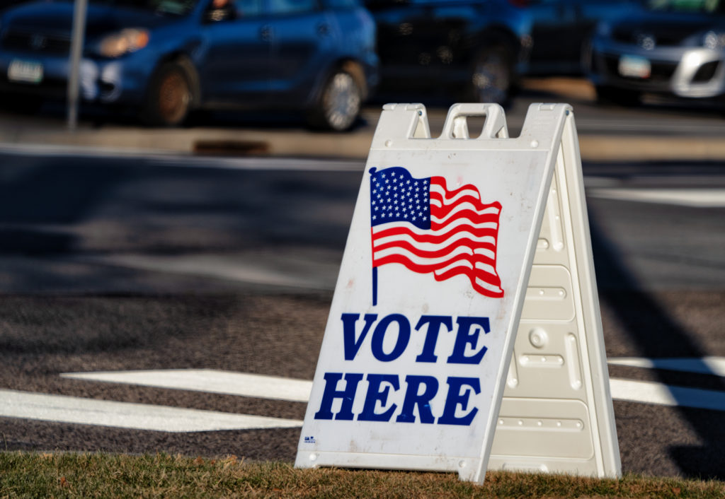 A white folding sign sits on a lawn. The American flag is drawn above "VOTE HERE" on the sign. A crosswalk and road are behind the sign.