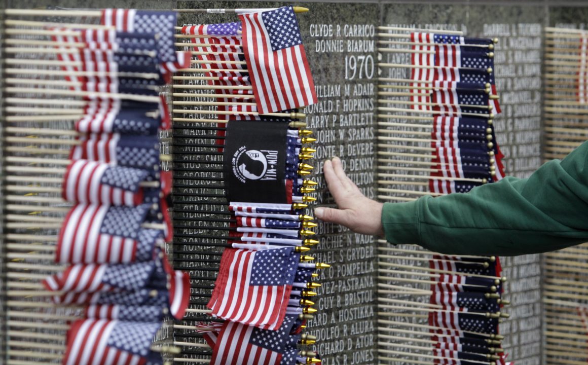 A hand rests against a memorial which is decorated by American flags and a POW/MIA flag.