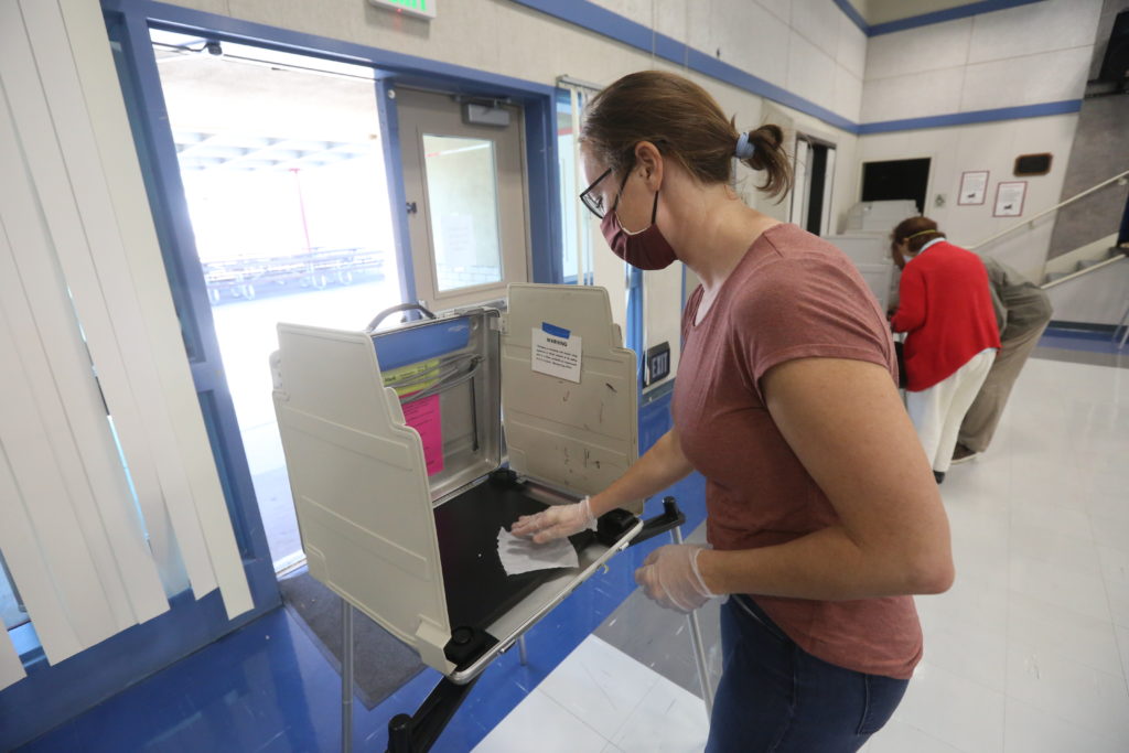 A woman in a mask uses a sanitizing wipe on a poll both.