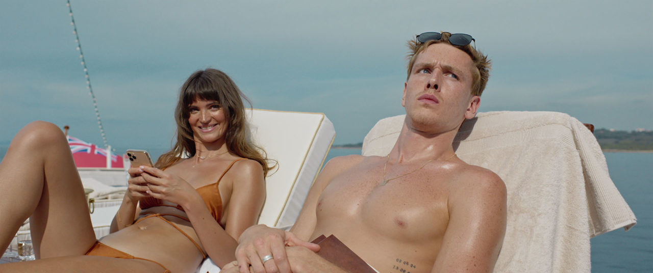 A bikini clad woman with long blonde hair sits on a lounge chair smiling next to a shirtless blonde-haired man who looks perturbed with the ocean and the blue sky visible behind them.