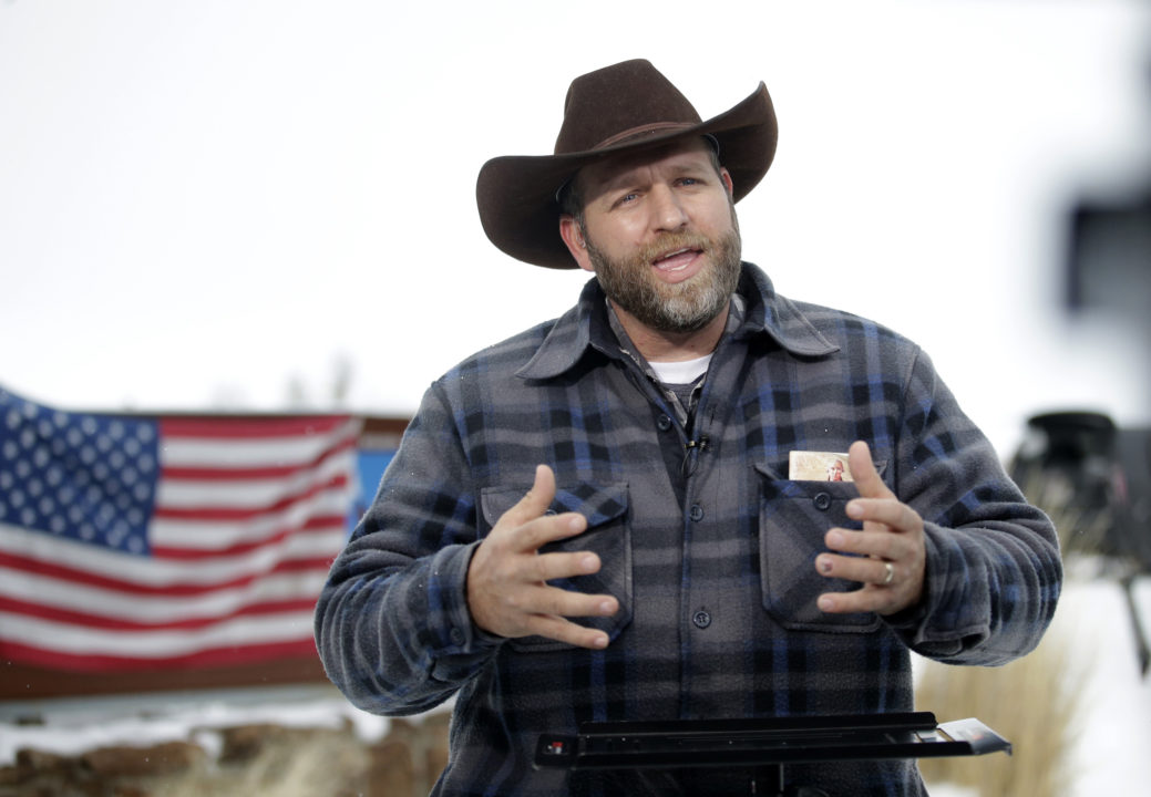 Idaho gubernatorial candidate Ammon Bundy stands in front of an American flag wearing a brown cowboy hat and blue plaid button down shirt with his hands in the air as he speaks emphatically.