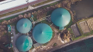 An aerial view of four massive bright green metallic biogas containers in the middle of a field of dirt and grass.