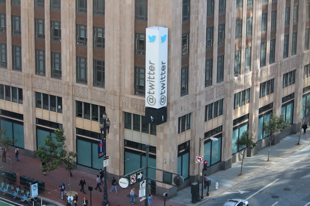 A large brown stone building sits on a city block in broad daylight with people passing by while a white and blue sign that says "Twitter" hangs from the building.