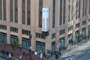 A large brown stone building sits on a city block in broad daylight with people passing by while a white and blue sign that says "Twitter" hangs from the building.