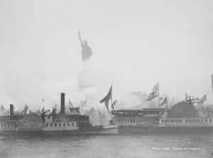 Steam boats sit in New York Harbor pushing steam from their chimneys in front of the Statue of Liberty in this black and white image from 1886.