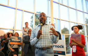 Senator Raphael Warnock addresses a crowd a crowd while holding a microphone and wearing a blue and white plaid shirt tucked into jeans.