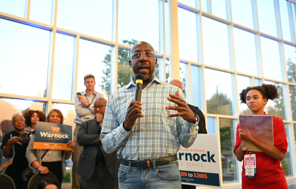 Senator Raphael Warnock addresses a crowd a crowd while holding a microphone and wearing a blue and white plaid shirt tucked into jeans.