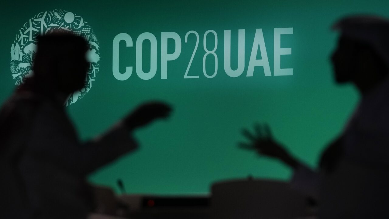 Two men sit silhouetted in front of a large projector screen with a green background that says "COP UAE".