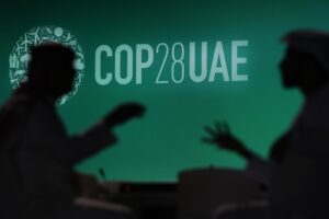 Two men sit silhouetted in front of a large projector screen with a green background that says "COP UAE".
