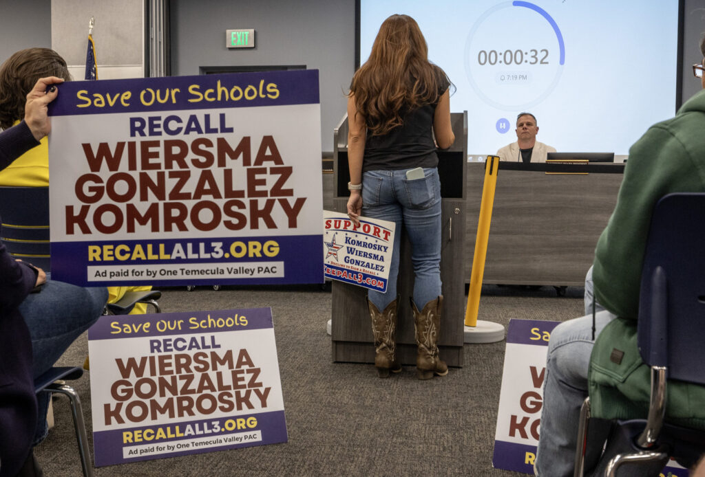 A woman with long brown hair stands at a podium with her back to the camera, speaking to a man sitting behind a desk as a person holds a sign behind the woman that says "Save Our Schools: Recall Wiersma, Gonzalez, Komrosky."